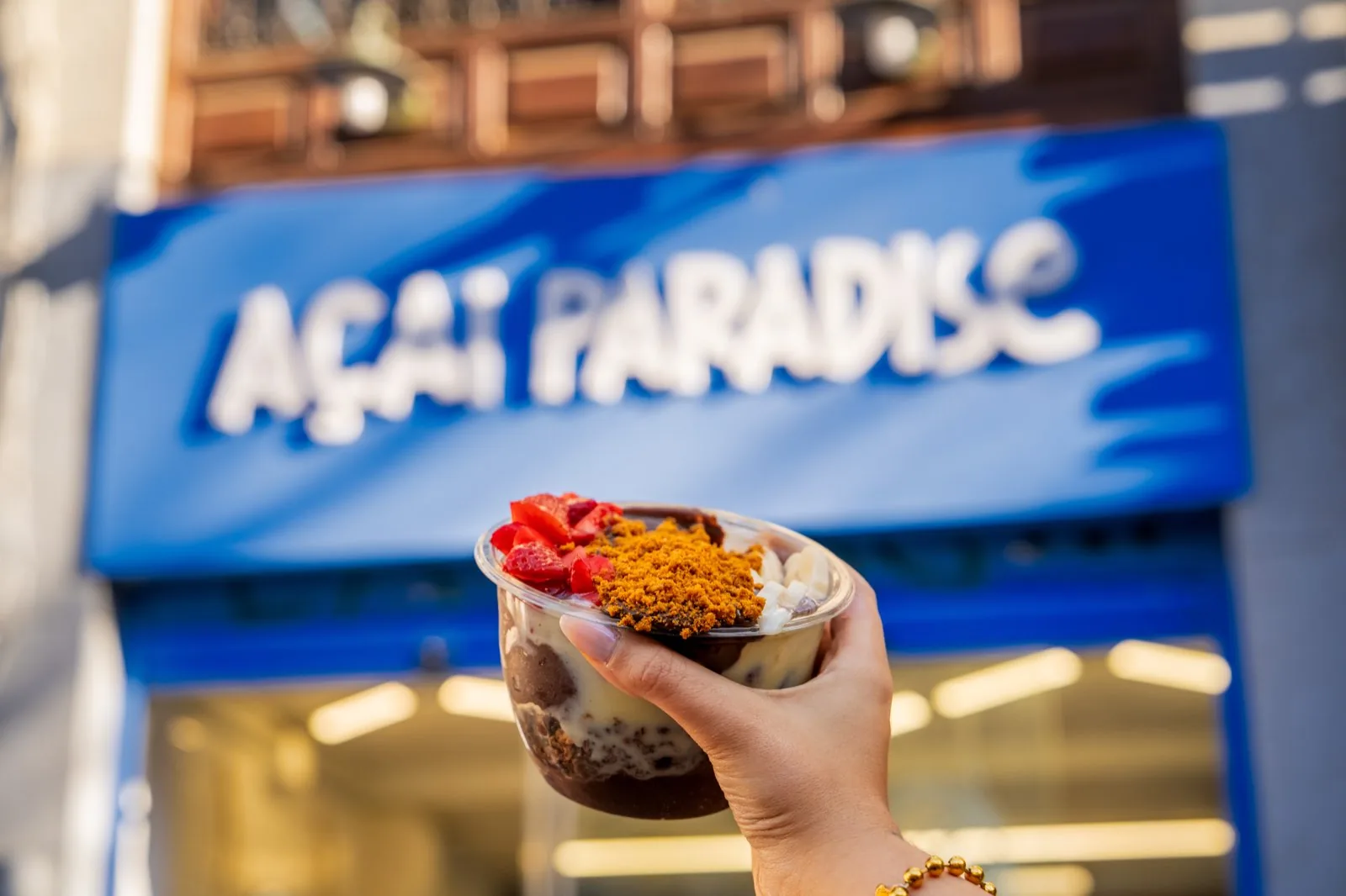 Acai bowl en la mano con el rótulo azul de Açaí Paradise Granada al fondo