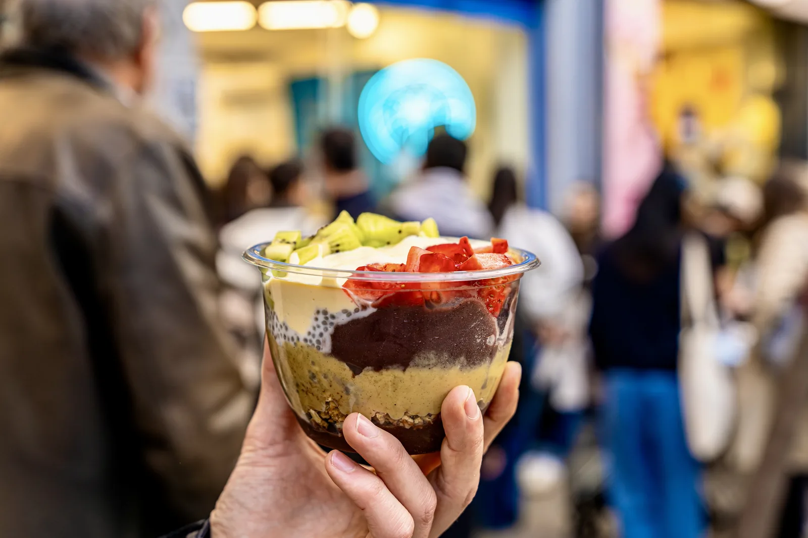 Bowl de açaí en la mano con clientes haciendo cola al fondo en Açaí Paradise